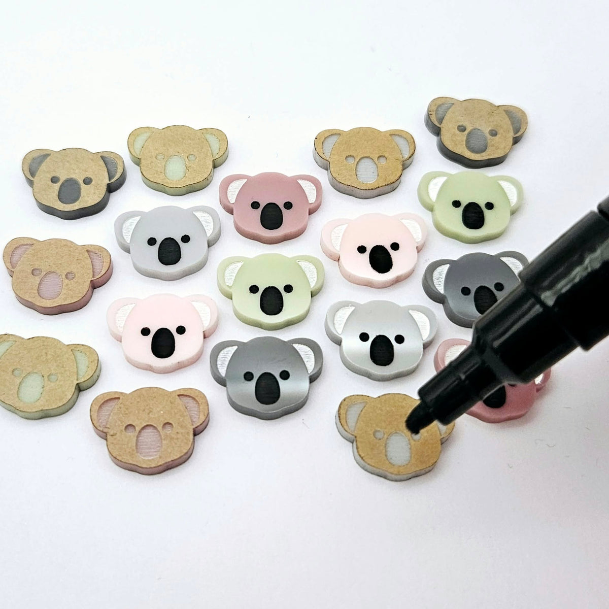 18mm solid acrylic koala stud earring blanks in Grey, Strawberry Milkshake, Dark Grey, Eucalyptus Earth and Dusty Rose, with five paint-filled pairs laid flat and additional unpainted koalas scattered around and an uncapped black Posca Pen.
