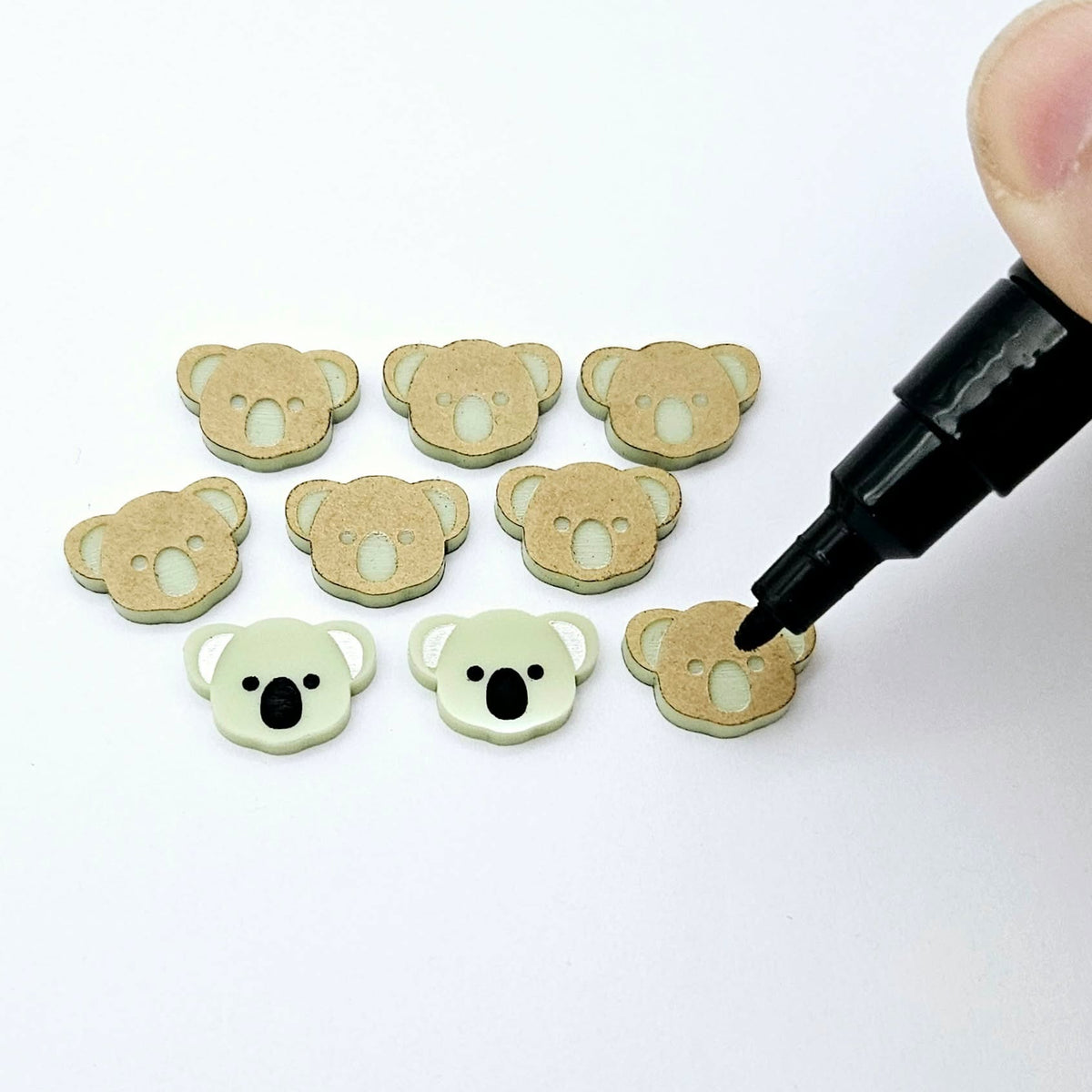 Solid Eucalyptus Green acrylic koala stud blanks showing one finished pair with protective paper removed and engraved details paint filled in black and white, alongside unpeeled blanks with protective paper still attached and a POSCA paint pen used for decorating.