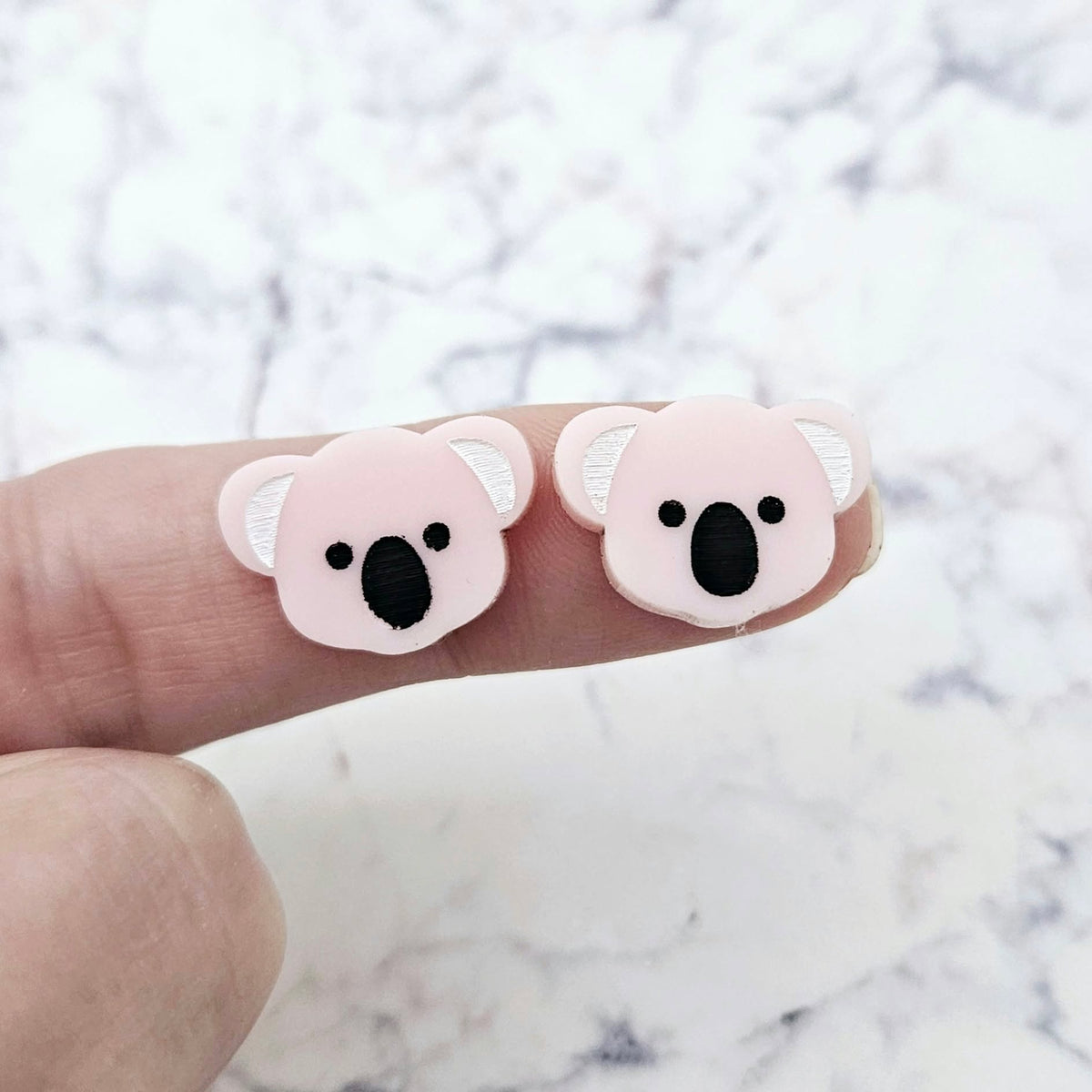 One pair of 18mm solid strawberry milkshake pink acrylic koala stud blanks displayed on a finger to show their petite size, featuring engraved eyes, nose and ears with black and white paint-filled details.