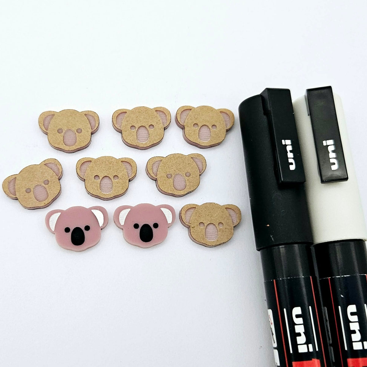 Solid Dusty Rose Pink acrylic koala stud blanks showing one finished pair with protective paper removed and engraved details paint filled in black and white, alongside unpeeled blanks with protective paper still attached and POSCA paint pens used for decorating.