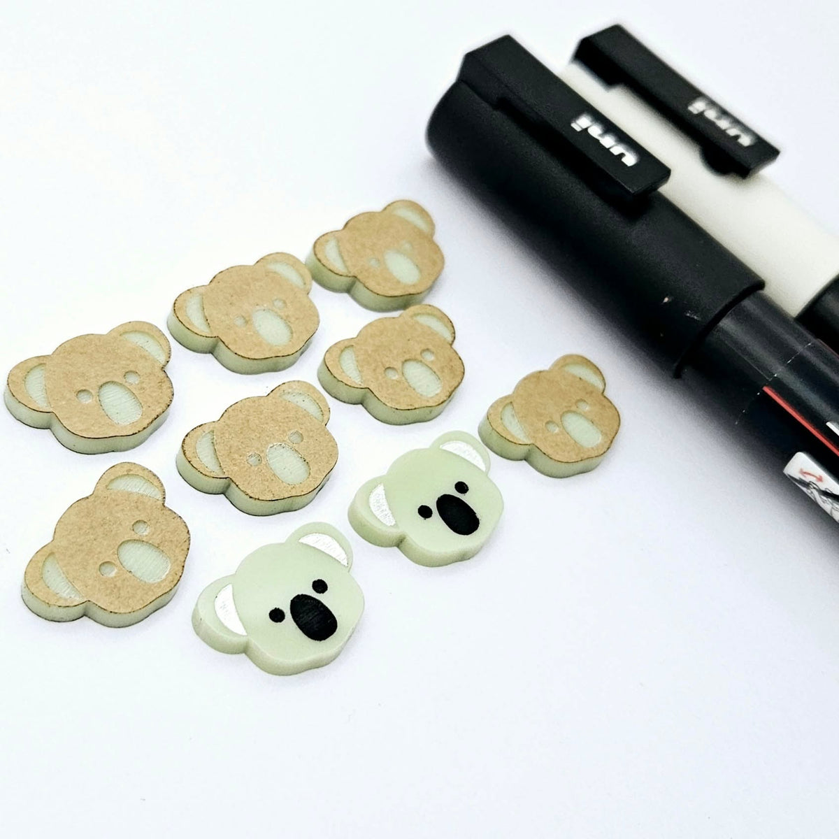 Solid Eucalyptus Green acrylic koala stud blanks showing one finished pair with protective paper removed and engraved details paint filled in black and white, alongside unpeeled blanks with protective paper still attached and POSCA paint pens used for decorating.