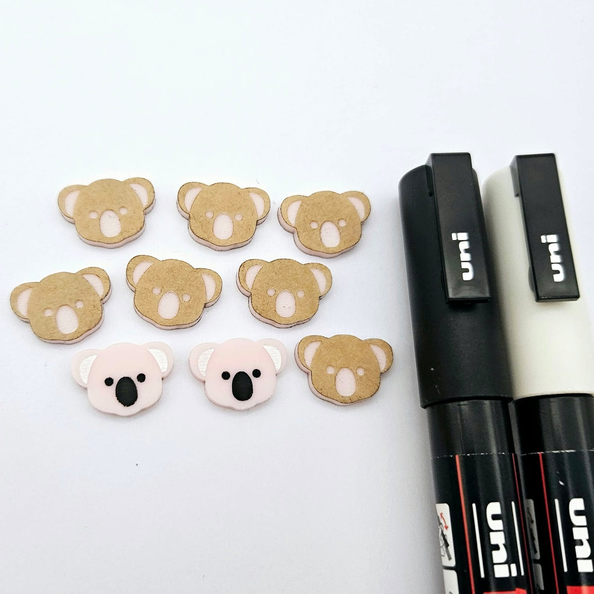 Solid Strawberry Milkshake Pink acrylic koala stud blanks showing one finished pair with protective paper removed and engraved details paint filled in black and white, alongside unpeeled blanks with protective paper still attached and POSCA paint pens used for decorating.