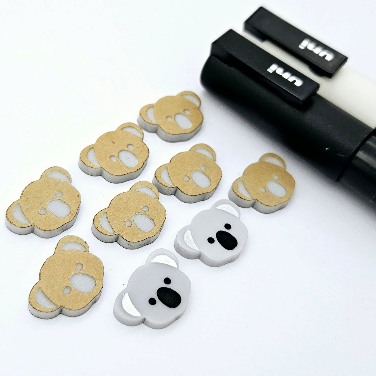 Solid Grey acrylic koala stud blanks showing one finished pair with protective paper removed and engraved details paint filled in black and white, alongside unpeeled blanks with protective paper still attached and POSCA paint pens used for decorating.