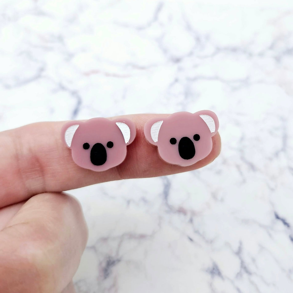 One pair of 18mm solid dusty rose pink acrylic koala stud blanks displayed on a finger to show their petite size, featuring engraved eyes, nose and ears with black and white paint-filled details.