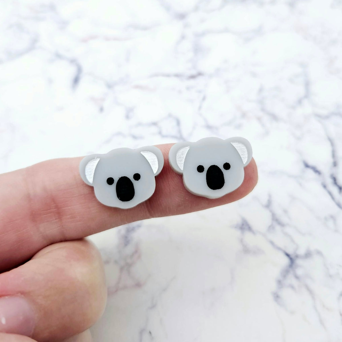One pair of 18mm solid grey acrylic koala stud blanks displayed on a finger to show their petite size, featuring engraved eyes, nose and ears with black and white paint-filled details.