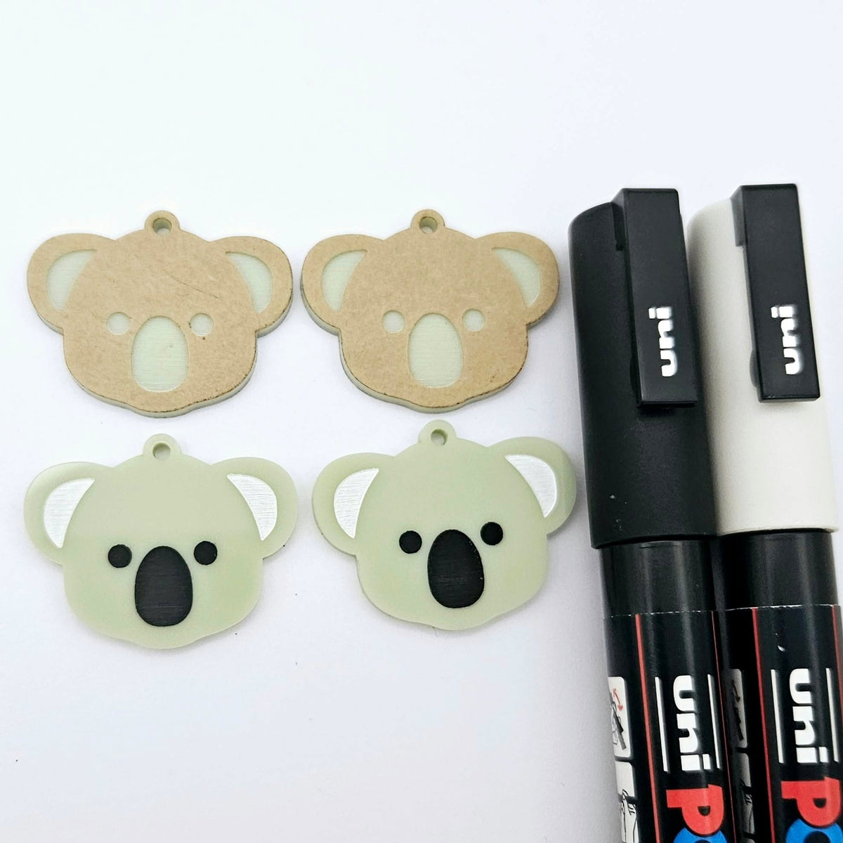 One pair of solid acrylic koala dangle earring blanks in Eucalyptus Green, laid flat with black and white paint-filled facial details, with another pair unpeeled koala blanks and POSCA paint pens.