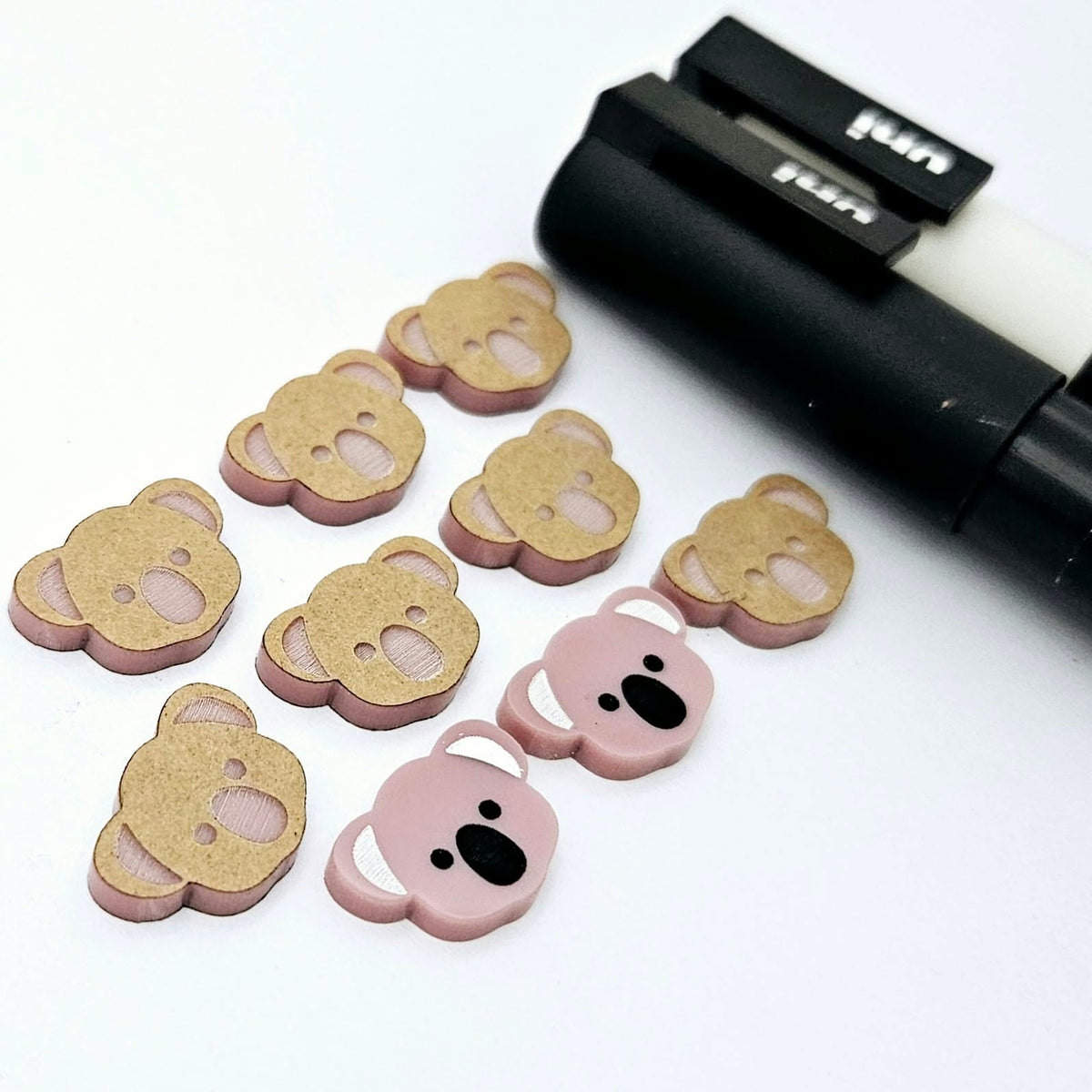 Solid Dusty Rose Pink acrylic koala stud blanks showing one finished pair with protective paper removed and engraved details paint filled in black and white, alongside unpeeled blanks with protective paper still attached and POSCA paint pens used for decorating.