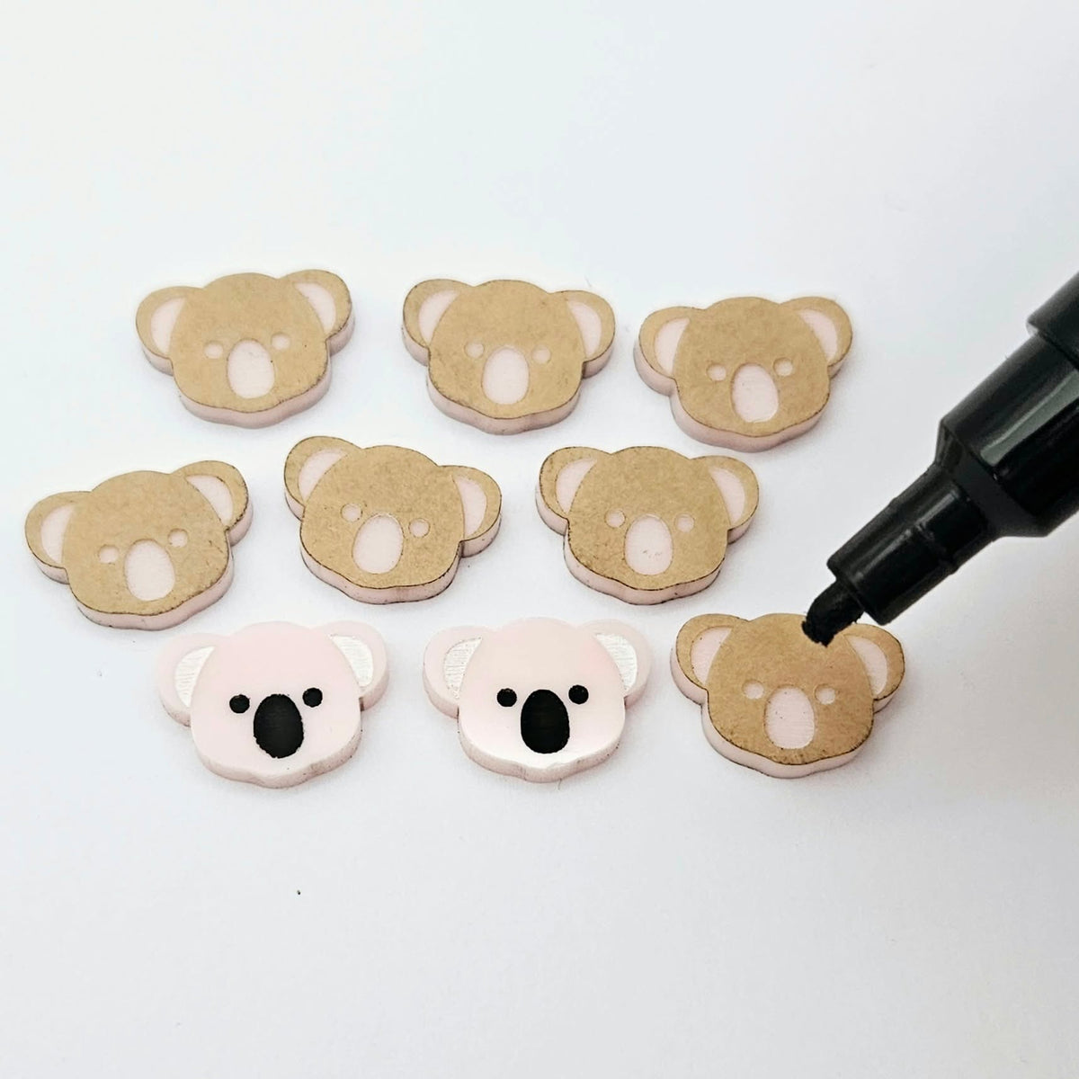 Solid strawberry milkshake pink acrylic koala stud blanks showing one finished pair with protective paper removed and engraved details paint filled in black and white, alongside unpeeled blanks with protective paper still attached and a POSCA paint pen used for decorating.