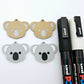 One pair of solid acrylic koala dangle earring blanks in grey, laid flat with black and white paint-filled facial details, with another pair unpeeled koala blanks and POSCA paint pens.