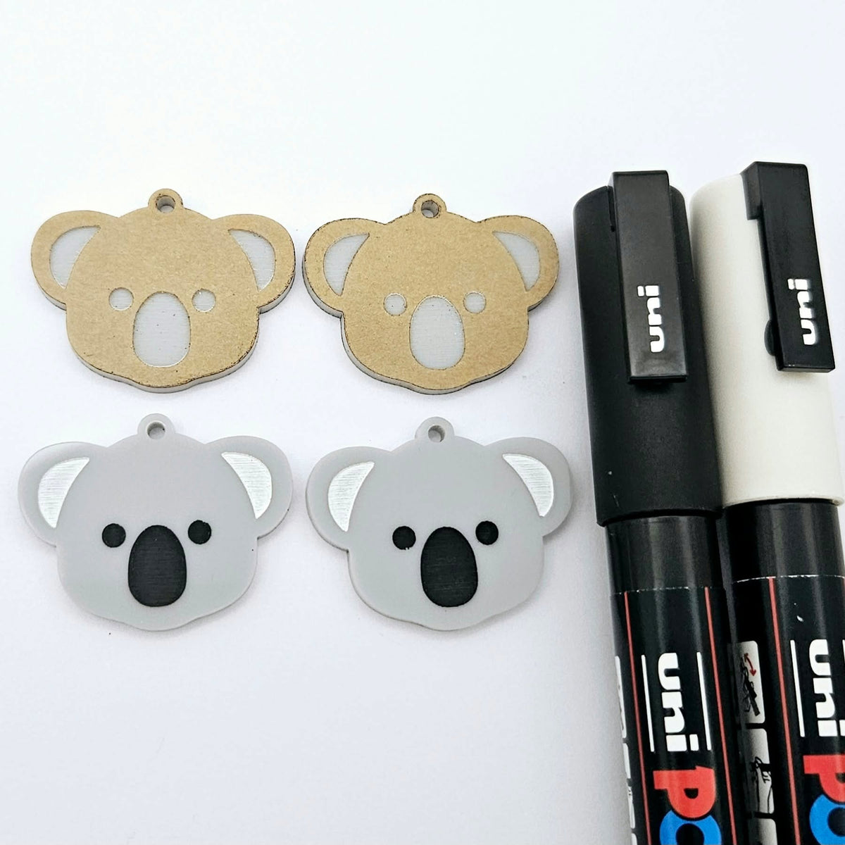 One pair of solid acrylic koala dangle earring blanks in grey, laid flat with black and white paint-filled facial details, with another pair unpeeled koala blanks and POSCA paint pens.
