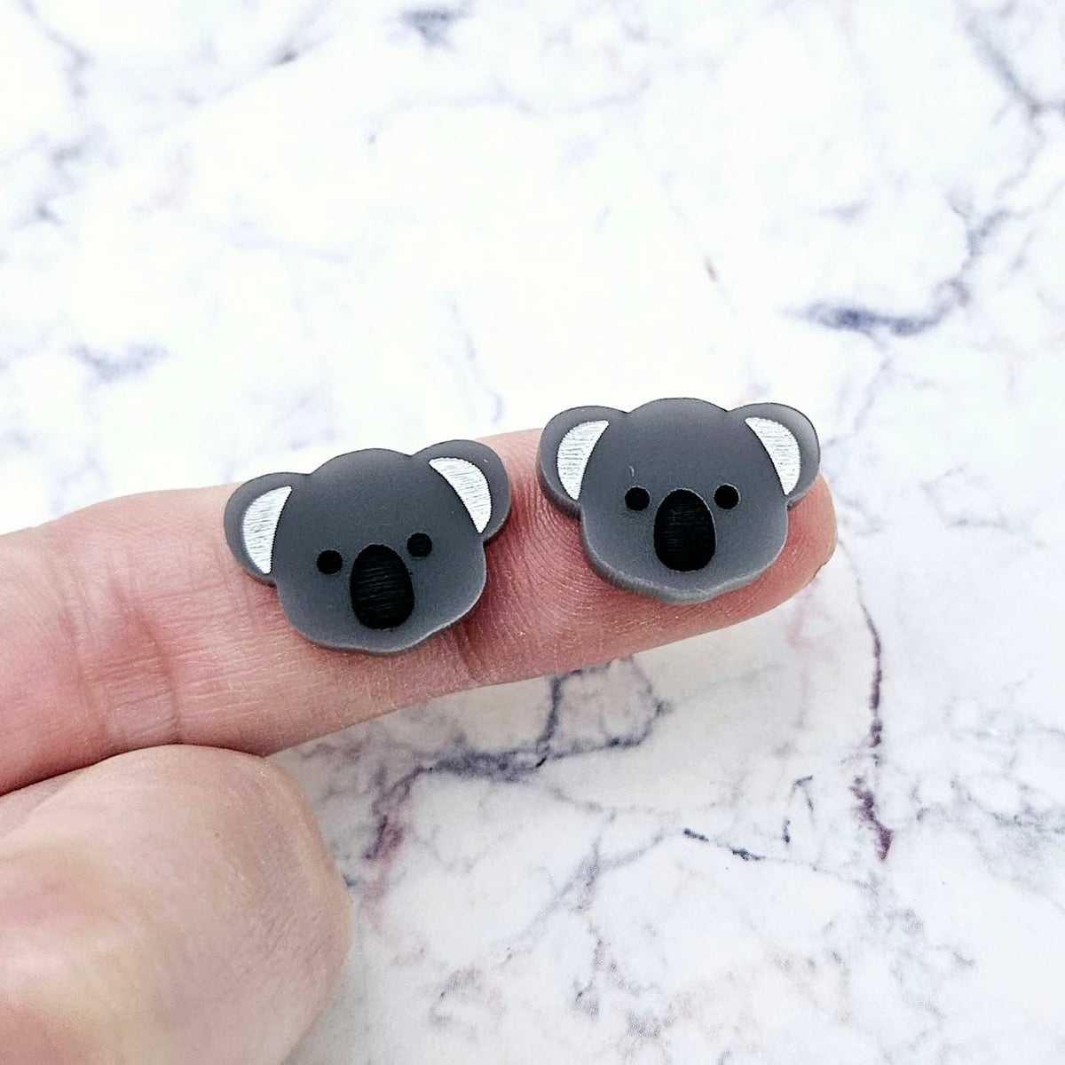 One pair of 18mm solid charcoal grey acrylic koala stud blanks displayed on a finger to show their petite size, featuring engraved eyes, nose and ears with black and white paint-filled details.