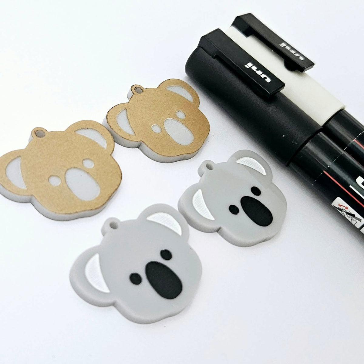 One pair of solid acrylic koala dangle earring blanks in grey, laid flat with black and white paint-filled facial details, with another pair unpeeled koala blanks and POSCA paint pens.