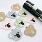 Solid acrylic koala dangle earring blanks in Grey, Strawberry Milkshake and Eucalyptus Green, laid flat with black and white paint-filled facial details, surrounded by unpeeled koala blanks and POSCA paint pens.