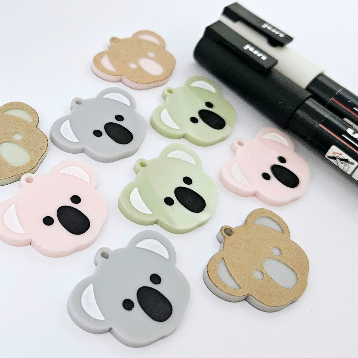 Solid acrylic koala dangle earring blanks in Grey, Strawberry Milkshake and Eucalyptus Green, laid flat with black and white paint-filled facial details, surrounded by unpeeled koala blanks and POSCA paint pens.