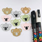 Solid acrylic koala dangle earring blanks in Grey, Strawberry Milkshake and Eucalyptus Green, laid flat with black and white paint-filled facial details, surrounded by unpeeled koala blanks and POSCA paint pens.