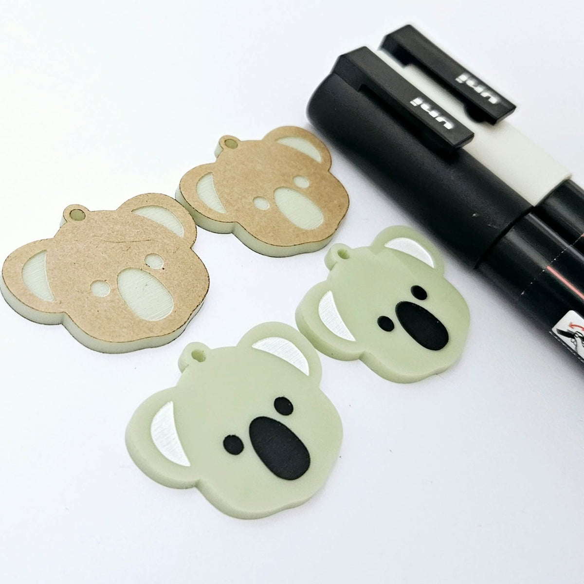 One pair of solid acrylic koala dangle earring blanks in Eucalyptus Green, laid flat with black and white paint-filled facial details, with another pair unpeeled koala blanks and POSCA paint pens.