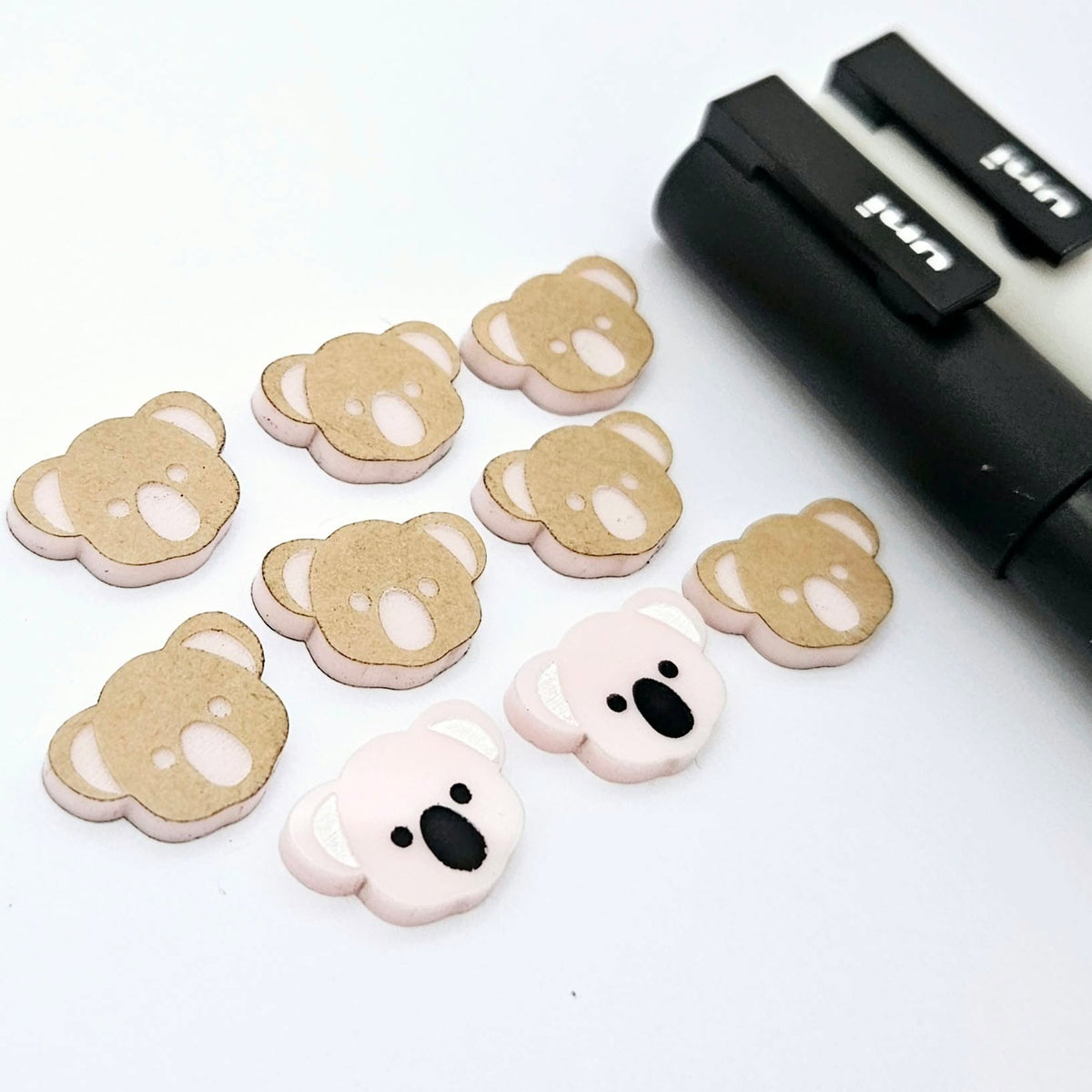 Solid Strawberry Milkshake Pink acrylic koala stud blanks showing one finished pair with protective paper removed and engraved details paint filled in black and white, alongside unpeeled blanks with protective paper still attached and POSCA paint pens used for decorating.
