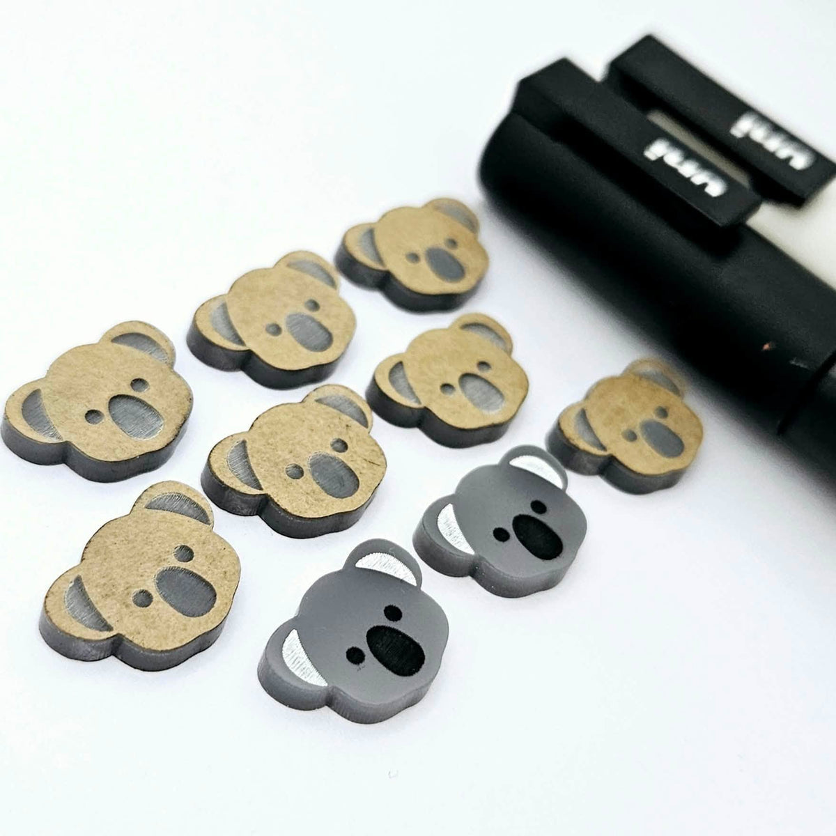 Solid charcoal grey acrylic koala stud blanks showing one finished pair with protective paper removed and engraved details paint filled in black and white, alongside unpeeled blanks with protective paper still attached and POSCA paint pens used for decorating.
