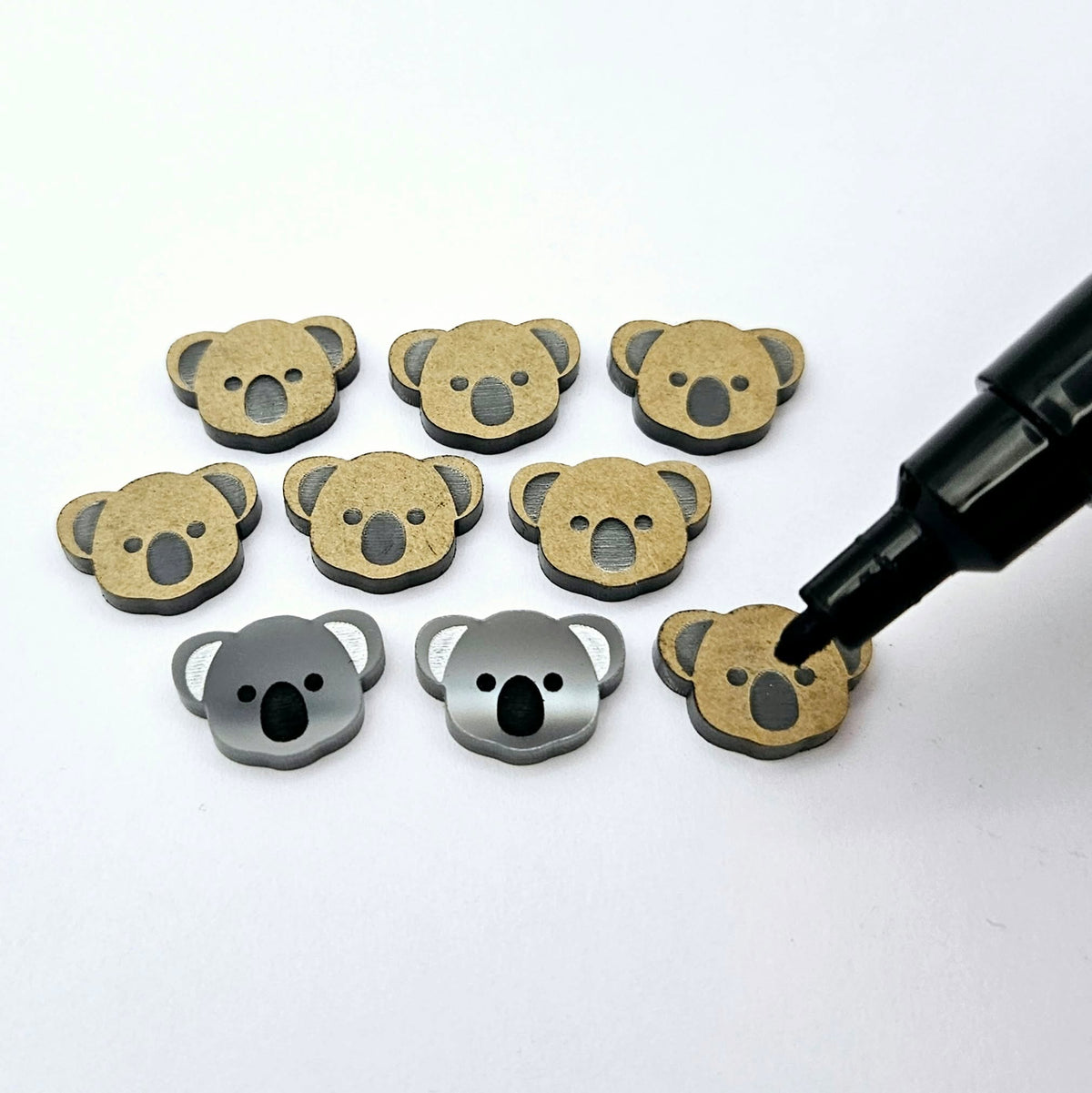 Solid charcoal grey acrylic koala stud blanks showing one finished pair with protective paper removed and engraved details paint filled in black and white, alongside unpeeled blanks with protective paper still attached and a POSCA paint pen used for decorating.