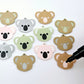 Solid acrylic koala dangle earring blanks in Grey, Strawberry Milkshake and Eucalyptus Green, laid flat with black and white paint-filled facial details, surrounded by unpeeled koala blanks and an uncapped black POSCA paint pen ready to paint.