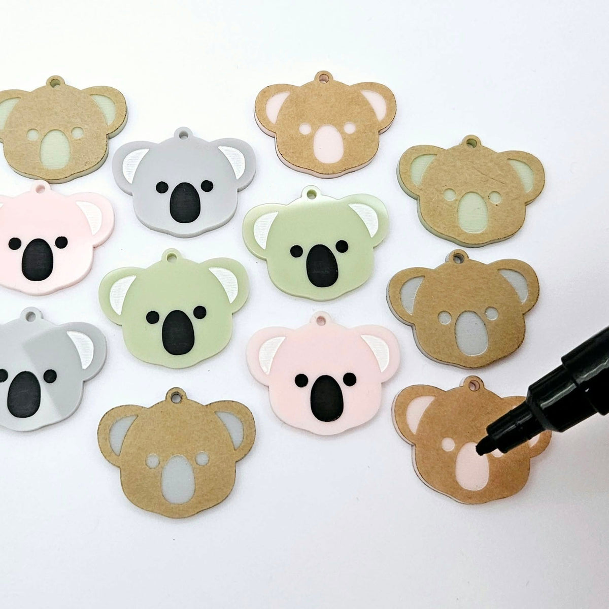 Solid acrylic koala dangle earring blanks in Grey, Strawberry Milkshake and Eucalyptus Green, laid flat with black and white paint-filled facial details, surrounded by unpeeled koala blanks and an uncapped black POSCA paint pen ready to paint.