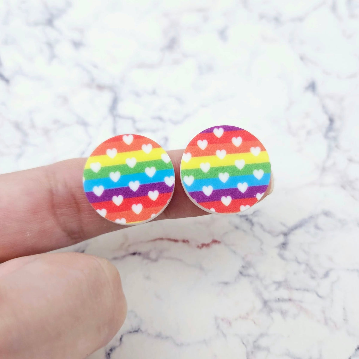 One pair of 20mm Rainbow Love UV printed acrylic stud earring blanks displayed on a finger to show their size, perfect for creating fun statement stud earrings.