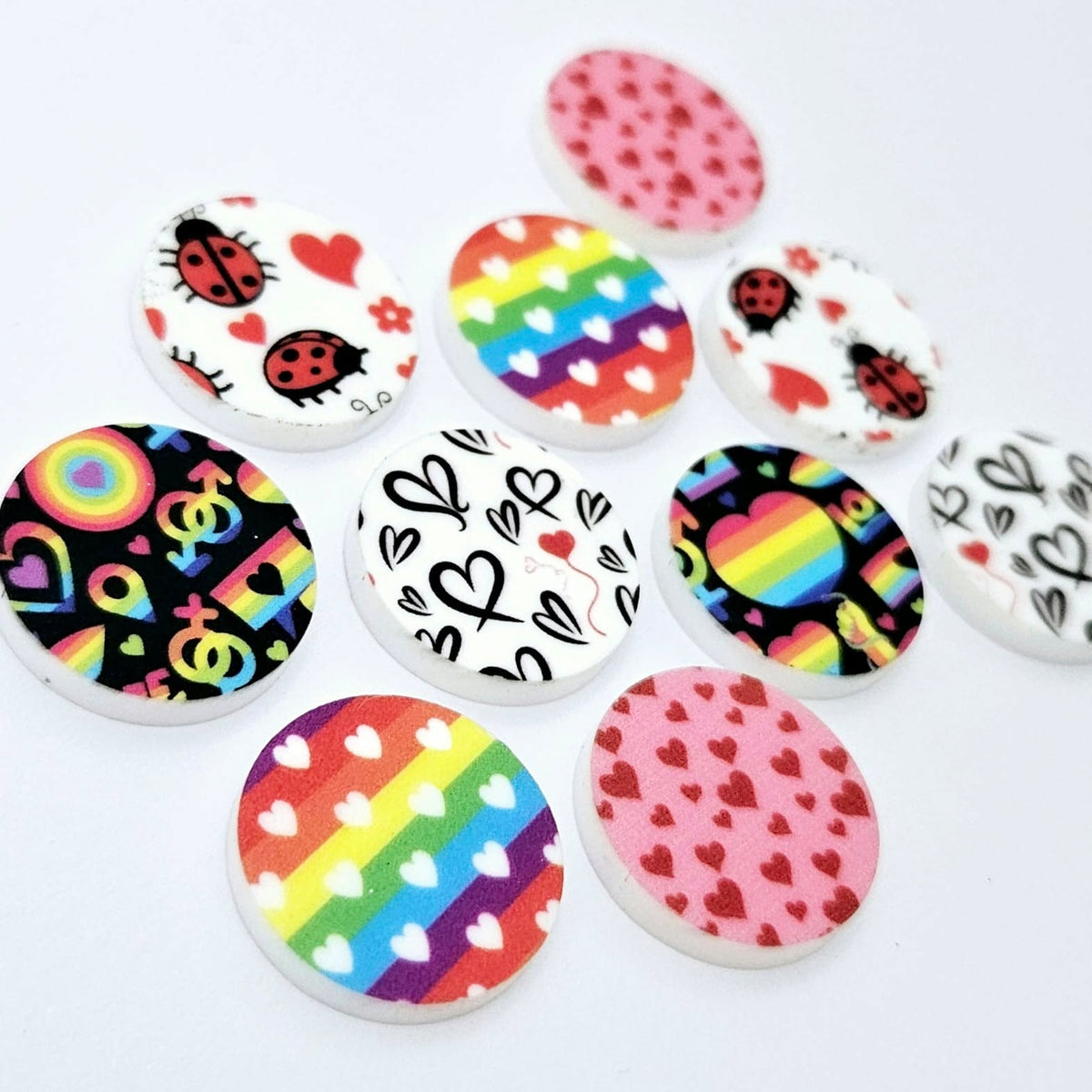 20mm UV printed Love Is Love acrylic stud earring blanks in five colourful designs: Neon Pride, Rainbow Love, Pink & Red Hearts, Black & Red Hearts and Ladybug Love, with all five pairs laid flat together.