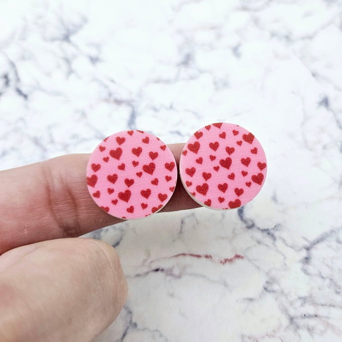 One pair of 20mm Pink & Red Hearts UV printed acrylic stud earring blanks displayed on a finger to show their size, perfect for creating fun statement stud earrings.