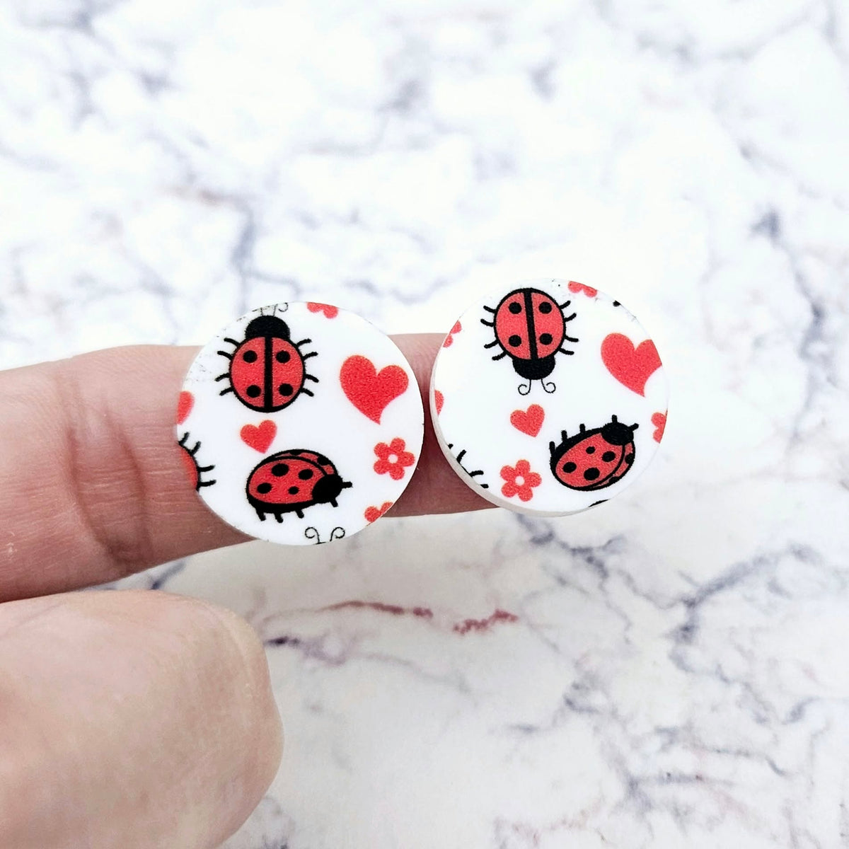 One pair of 20mm Ladybug Love UV printed acrylic stud earring blanks displayed on a finger to show their size, perfect for creating fun statement stud earrings.