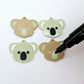 One pair of solid acrylic koala dangle earring blanks in Eucalyptus Green, laid flat with black and white paint-filled facial details, with another pair unpeeled koala blanks and a black uncapped posca paint pen.