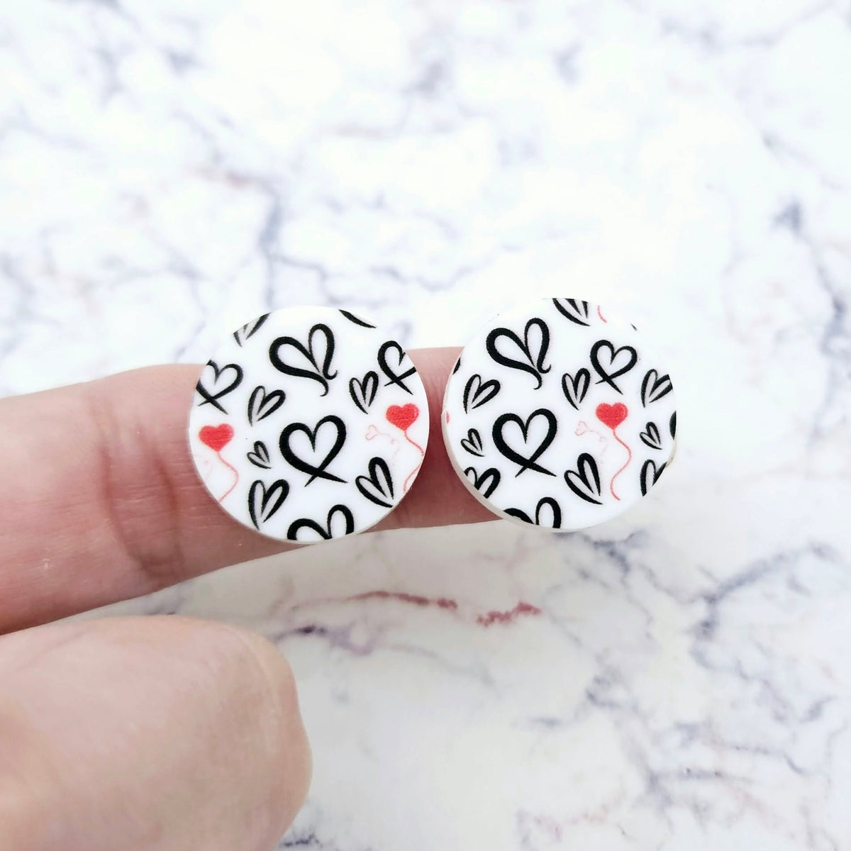 One pair of 20mm Black & Red Hearts UV printed acrylic stud earring blanks displayed on a finger to show their size, perfect for creating fun statement stud earrings.