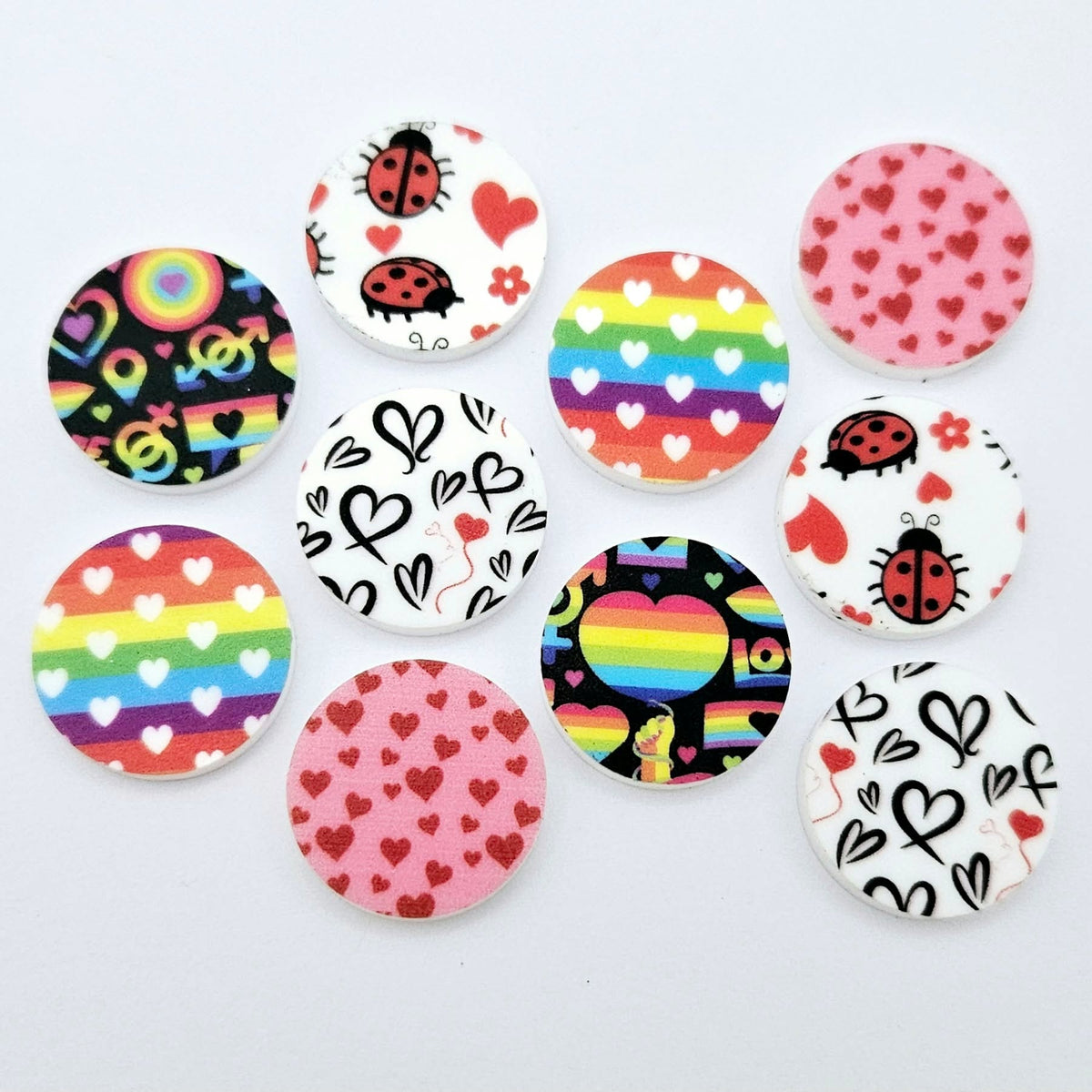 20mm UV printed Love Is Love acrylic stud earring blanks in five colourful designs: Neon Pride, Rainbow Love, Pink & Red Hearts, Black & Red Hearts and Ladybug Love, with all five pairs laid flat together.