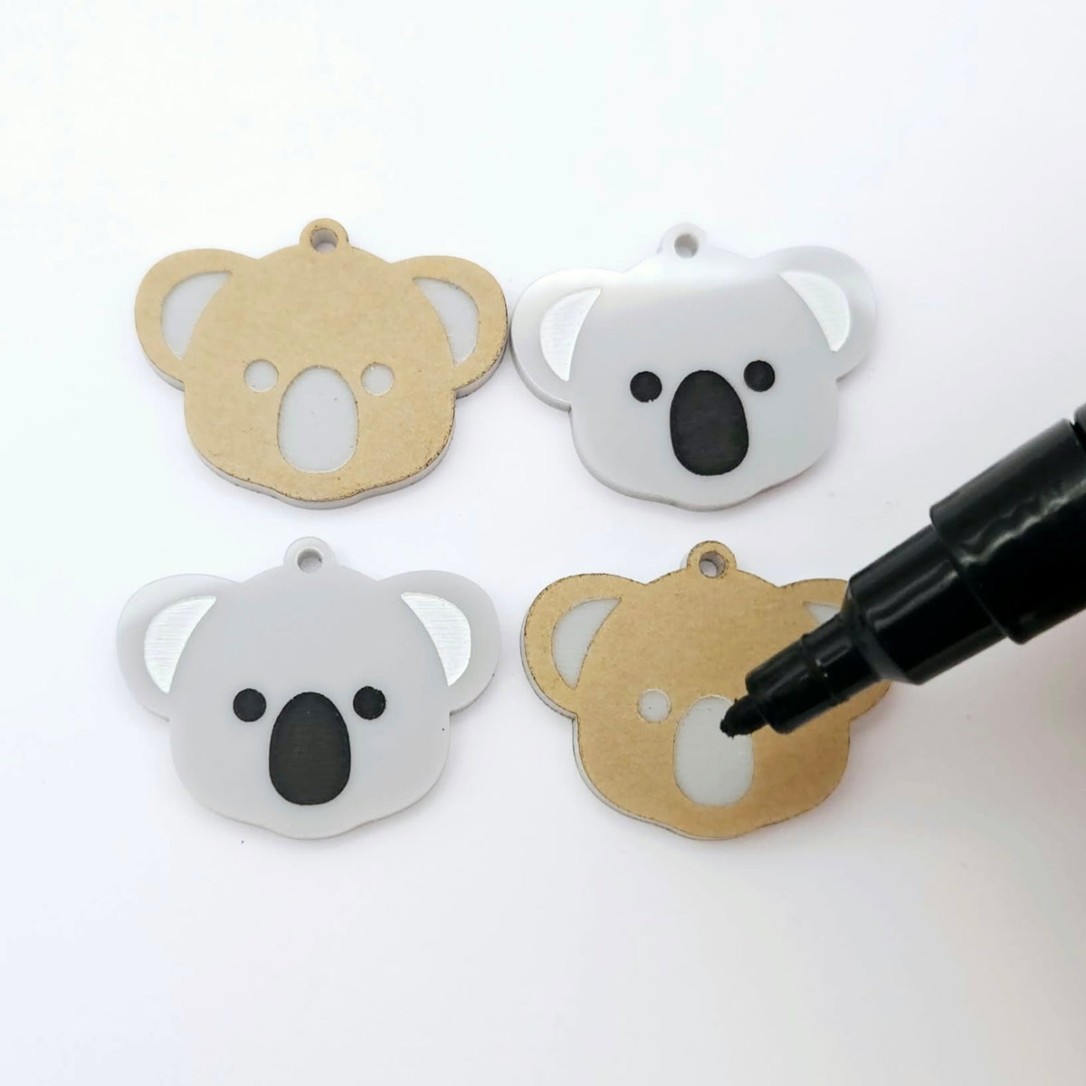 One pair of solid acrylic koala dangle earring blanks in grey, laid flat with black and white paint-filled facial details, with another pair unpeeled koala blanks and a black uncapped posca paint pen.